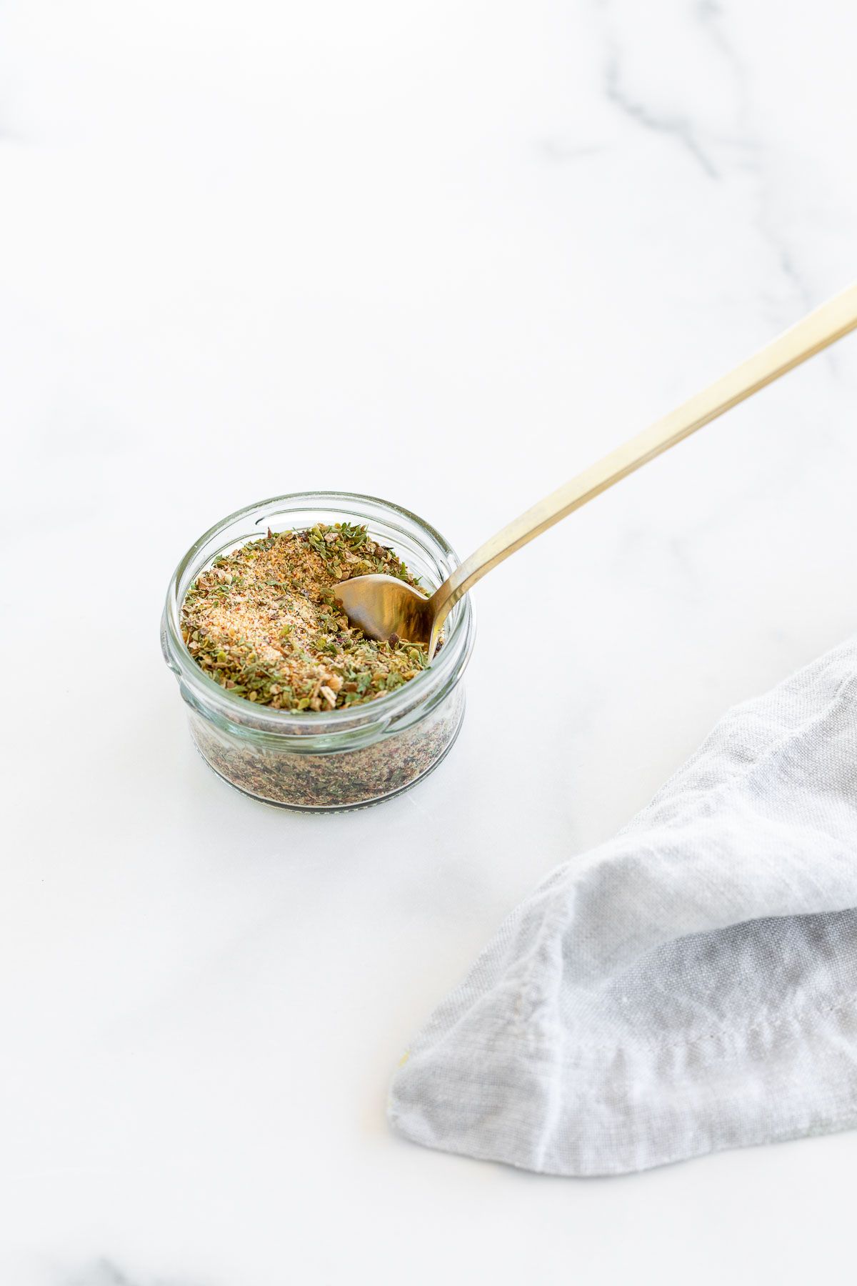 A small glass container filled with all purpose seasoning, gold spoon in the jar.