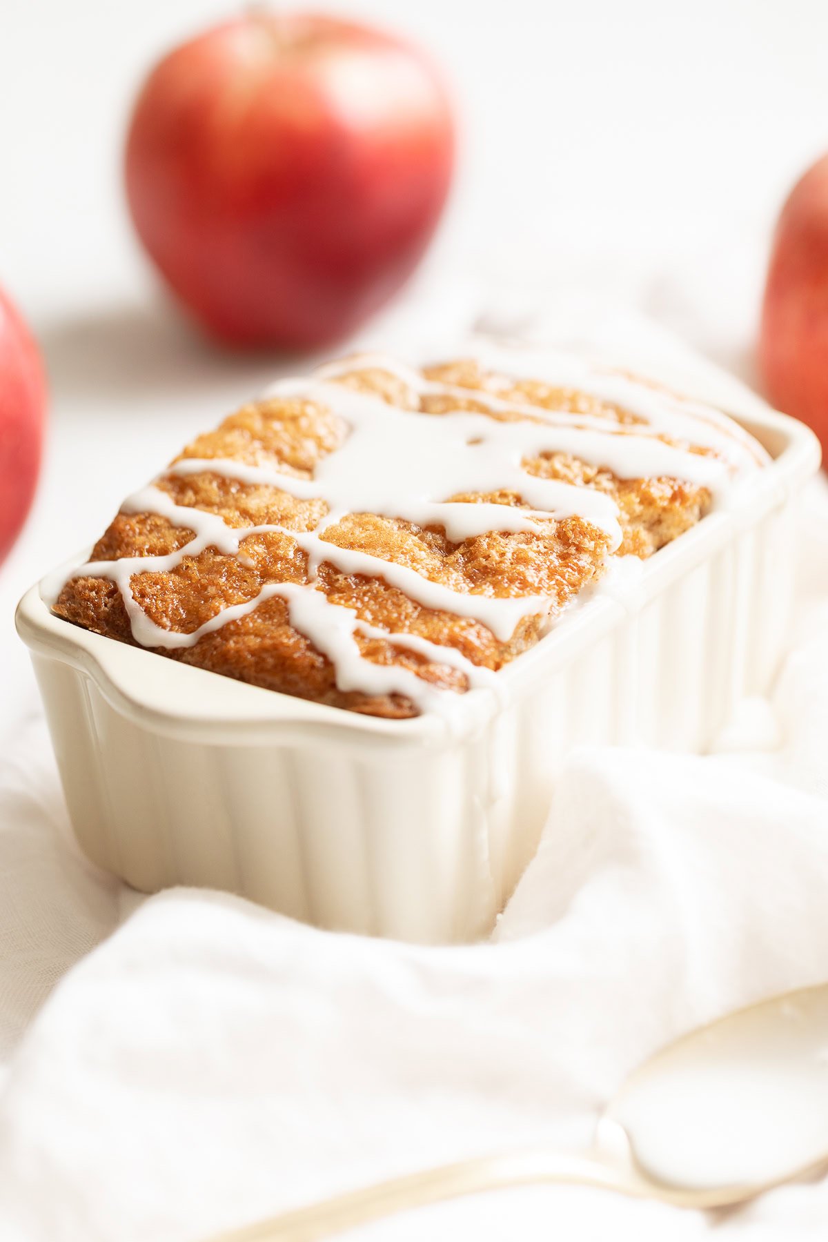 A small loaf pan of apple bread, topped with white icing and surrounded with apples and a linen napkin.