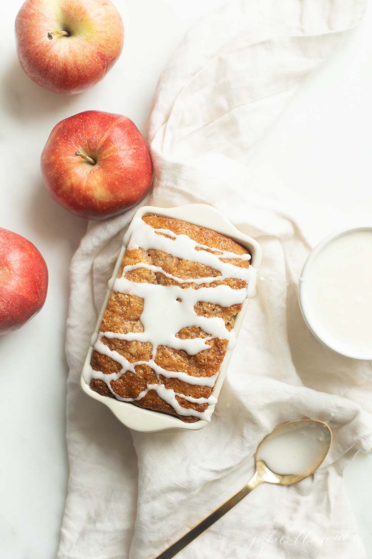 A small loaf pan of apple bread, topped with white icing and surrounded with apples and a linen napkin.