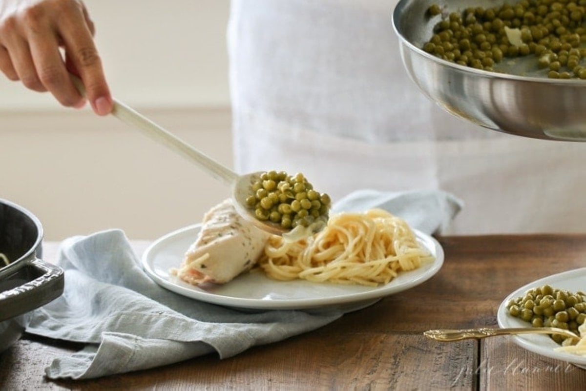 peas being spooned over a plate of pasta and prosciutto wrapped chicken