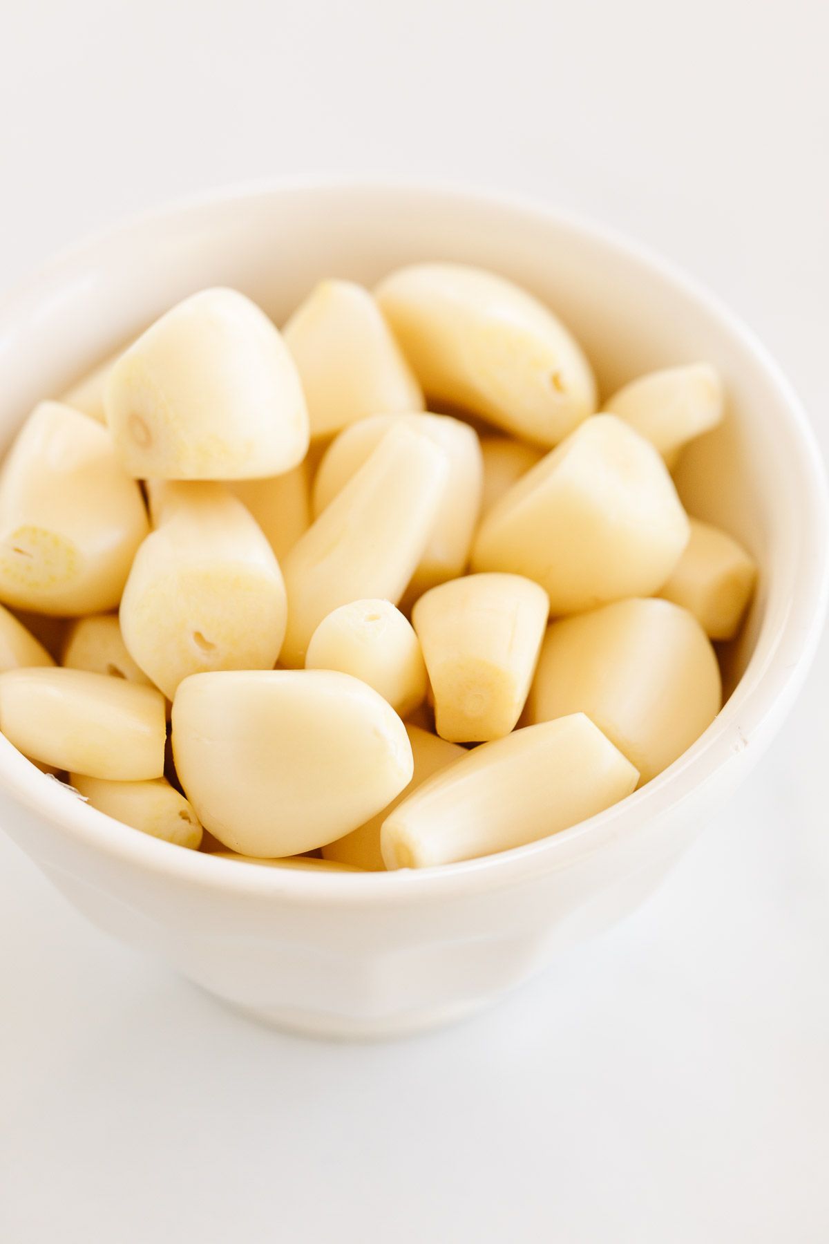 A white bowl full of freshly peeled garlic cloves, resting on a marble countertop