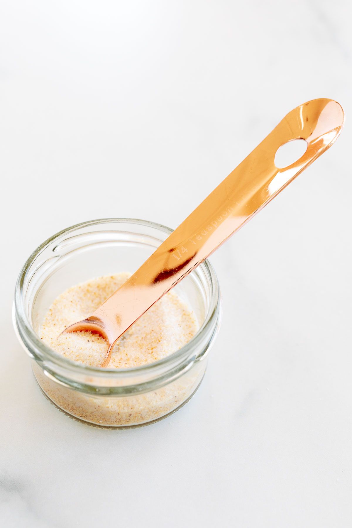 A small glass jar filled with homemade garlic salt, placed on a marble countertop. A copper measuring spoon is in the jar.