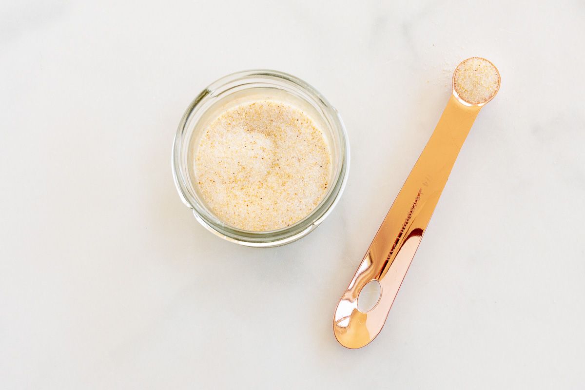 A small glass jar filled with homemade garlic salt, placed on a marble countertop. A copper measuring spoon is placed near the jar.