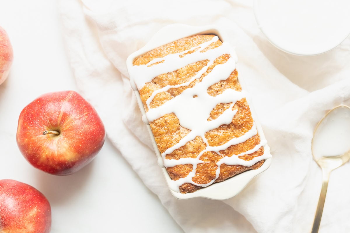 A delightful apple bread recipe features a loaf cake topped with white icing, perfectly placed in a dish. It's accompanied by two vibrant red apples, a glass of milk, and a spoon elegantly resting on a white cloth.