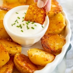 A hand dipping a Parmesan roasted potato into a white bowl of sour cream dip