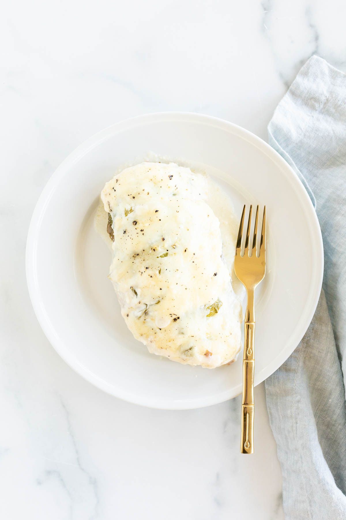 A baked chicken breast on a white plate, gold fork to the side.