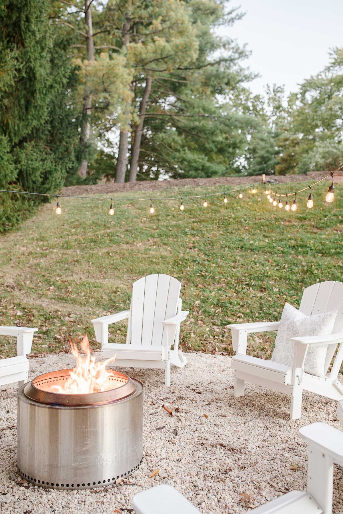 White Adirondack chairs, circling a Solo Stove on a DIY pea gravel fire pit patio.