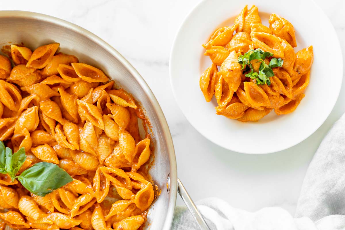 A white plate with a serving of Gigi Hadid pasta, a vodka pasta recipe. Gold fork on the side for serving, pan full of pasta on the side.