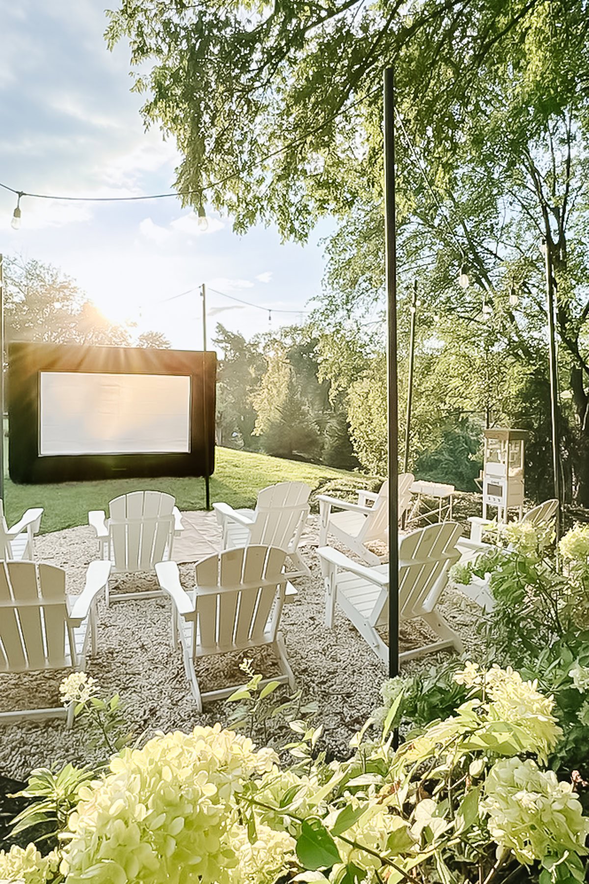 White Adirondack chairs are arranged on pea gravel in front of a large screen for an outdoor movie night. Surrounded by trees and twinkling string lights, the cozy setting is illuminated by the sun, with a nearby fire pit patio adding warmth and charm.