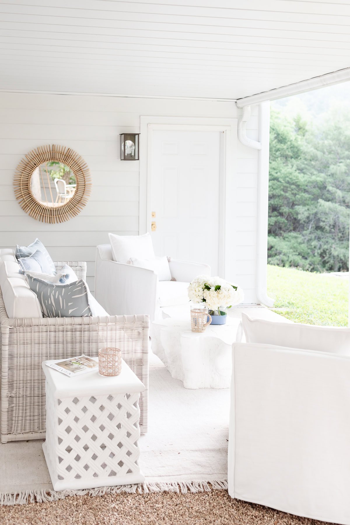 A covered porch hosts white wicker furniture with neutral cushions, complemented by a round mirror and decor. A small table and potted flowers add charm, while beyond the porch lies a cozy fire pit patio surrounded by lush greenery.