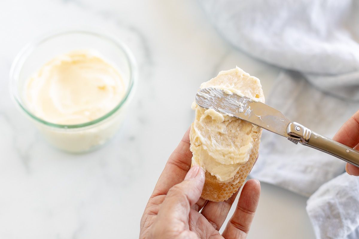 A hand holding a slice of French bread, spreading homemade butter on it.