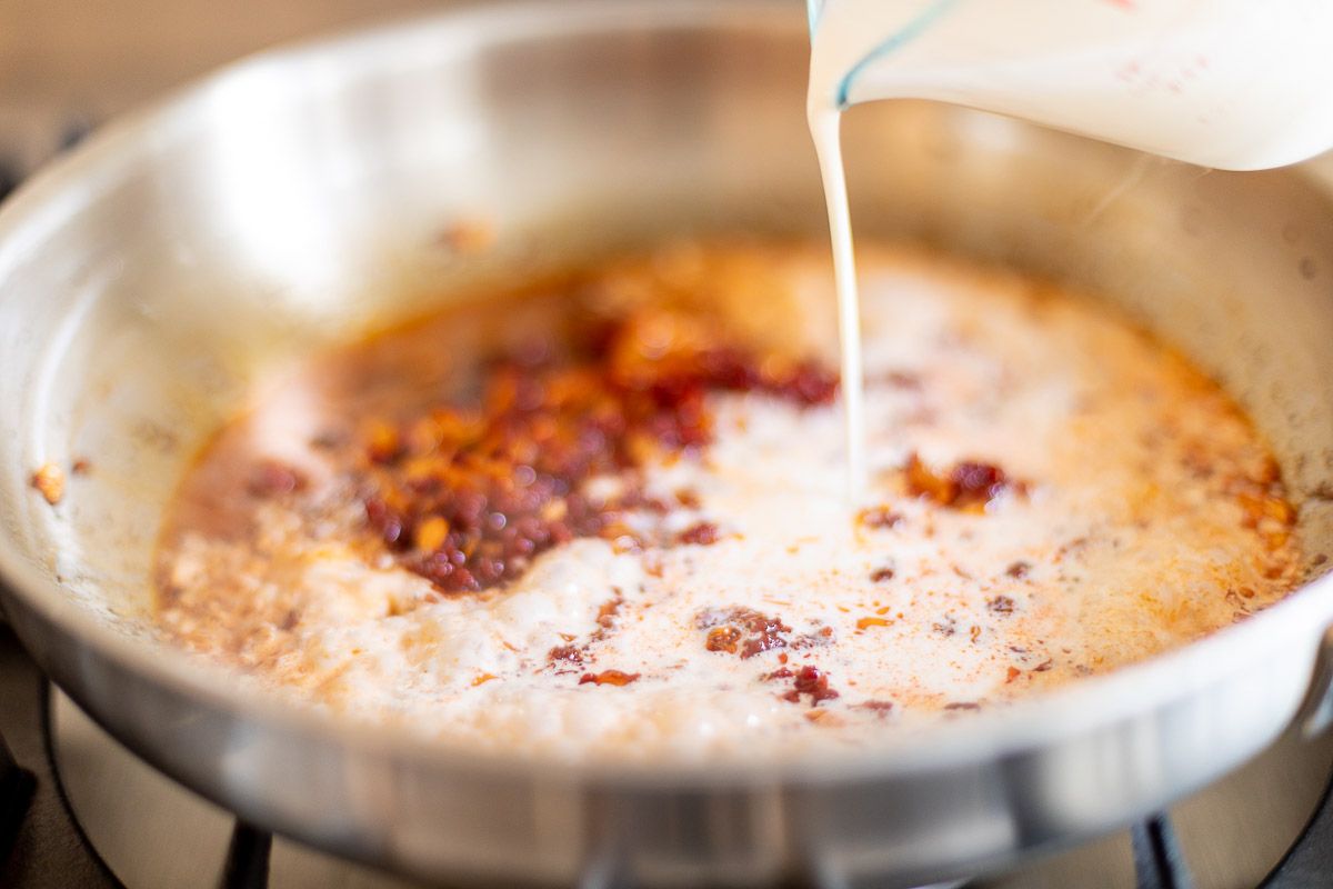 A glass pitcher of heavy whipping cream pouring into a pot of vodka sauce.