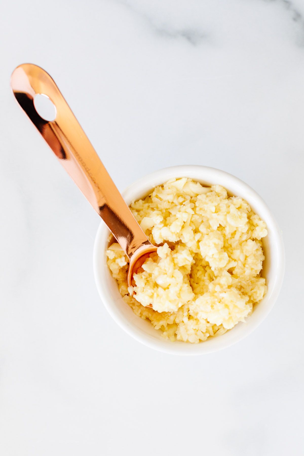 Minced garlic in a small white bowl, with a copper measuring spoon inside