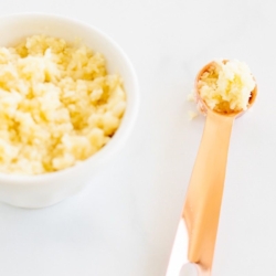 A small white bowl of minced garlic, with a copper teaspoon full of minced garlic next to it.