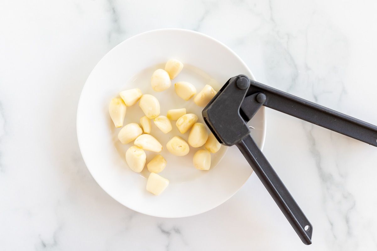 A hand holding a garlic press, mincing garlic over a white plate full of garlic cloves.