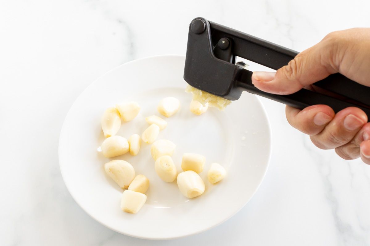 A hand holding a garlic press, mincing garlic over a white plate full of garlic cloves.