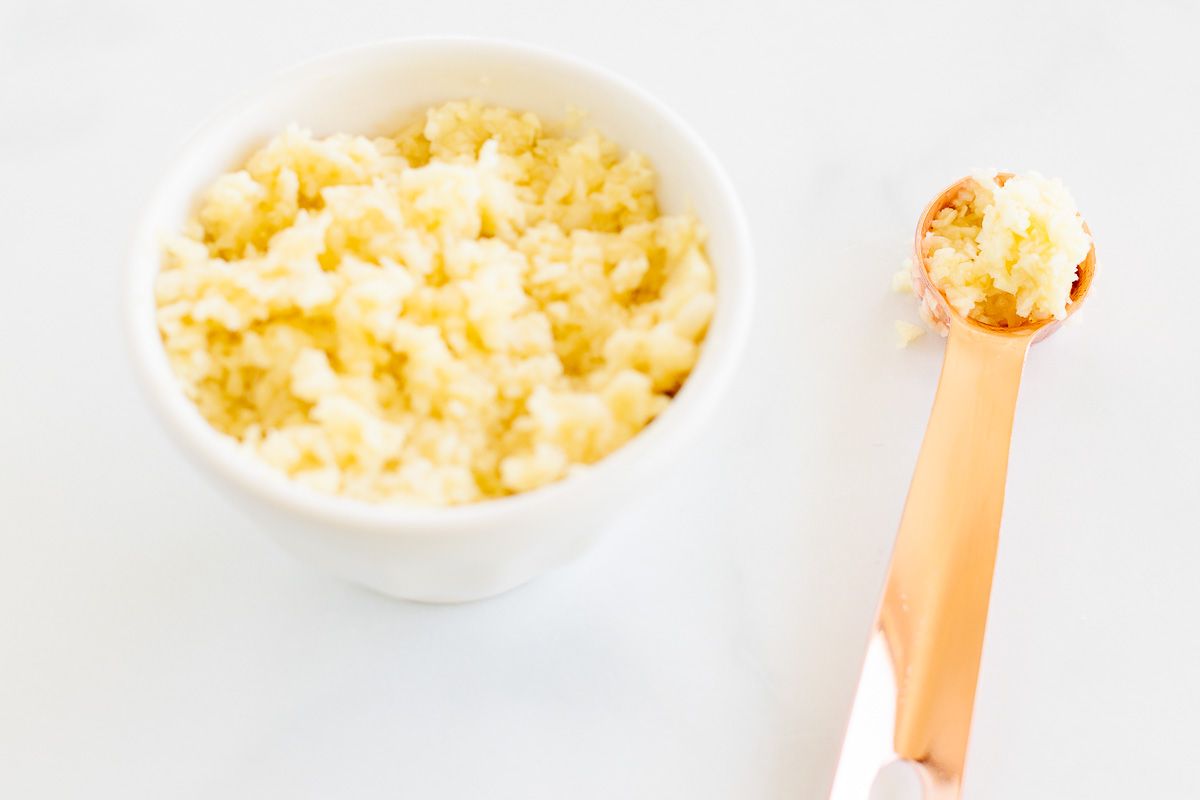 A small white bowl of minced garlic, with a copper teaspoon full of minced garlic next to it.