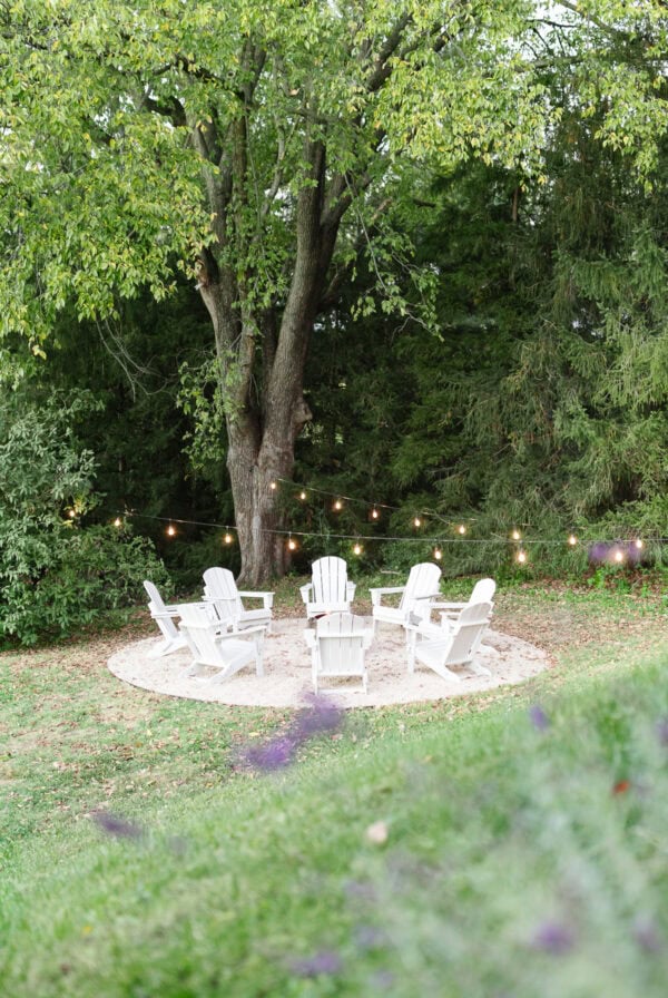Six white Adirondack chairs encircle a fire pit on a pea gravel patio under string lights, set on a round area surrounded by green grass and trees.