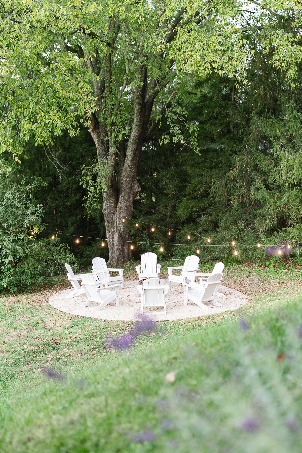 Six white Adirondack chairs encircle a fire pit on a pea gravel patio under string lights, set on a round area surrounded by green grass and trees.