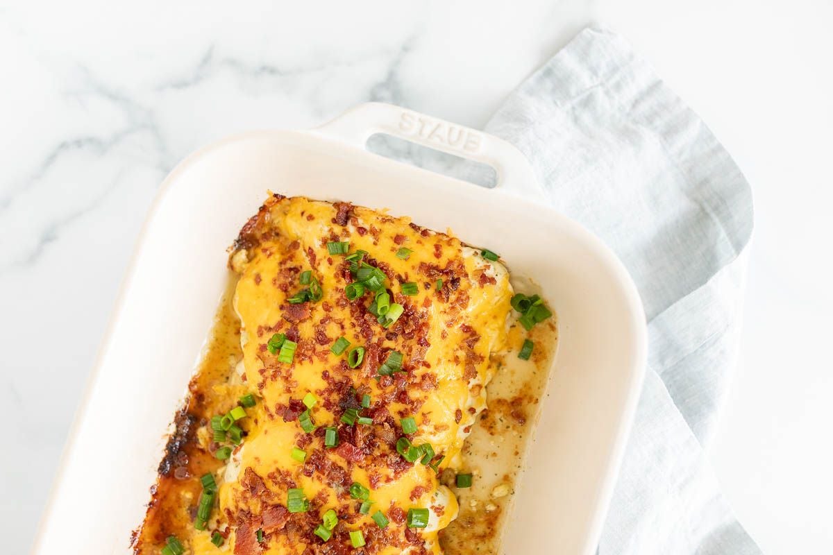 A white baking dish with cheesy crack chicken, topped with green onions and chopped bacon.