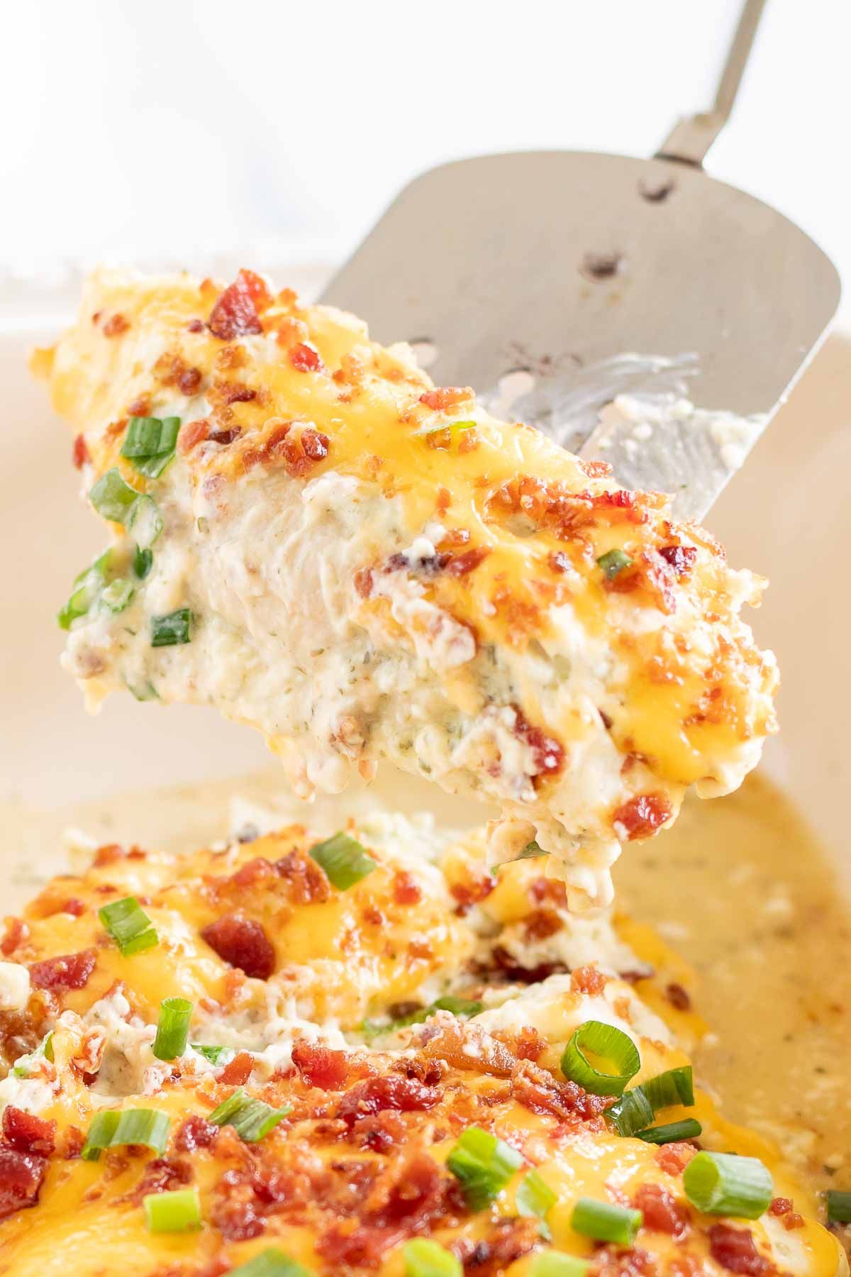 Baked crack chicken breasts in a white baking dish, with a silver spatula lifting a serving out.