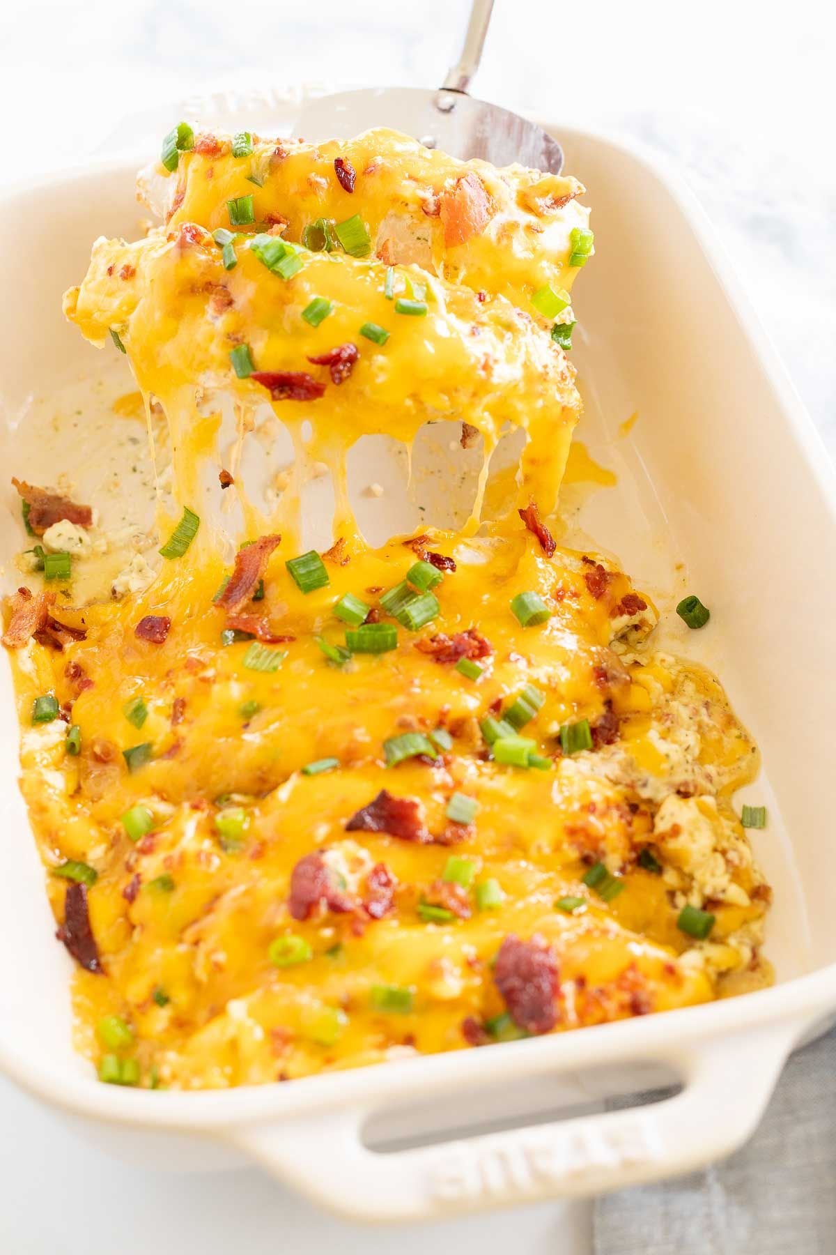 Baked crack chicken breasts in a white baking dish, with a silver spatula lifting a serving out. 