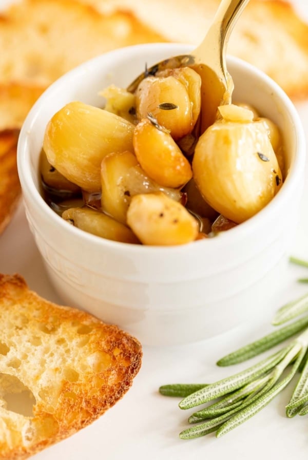 A white bowl full of garlic confit, surrounded by crostini and fresh rosemary.