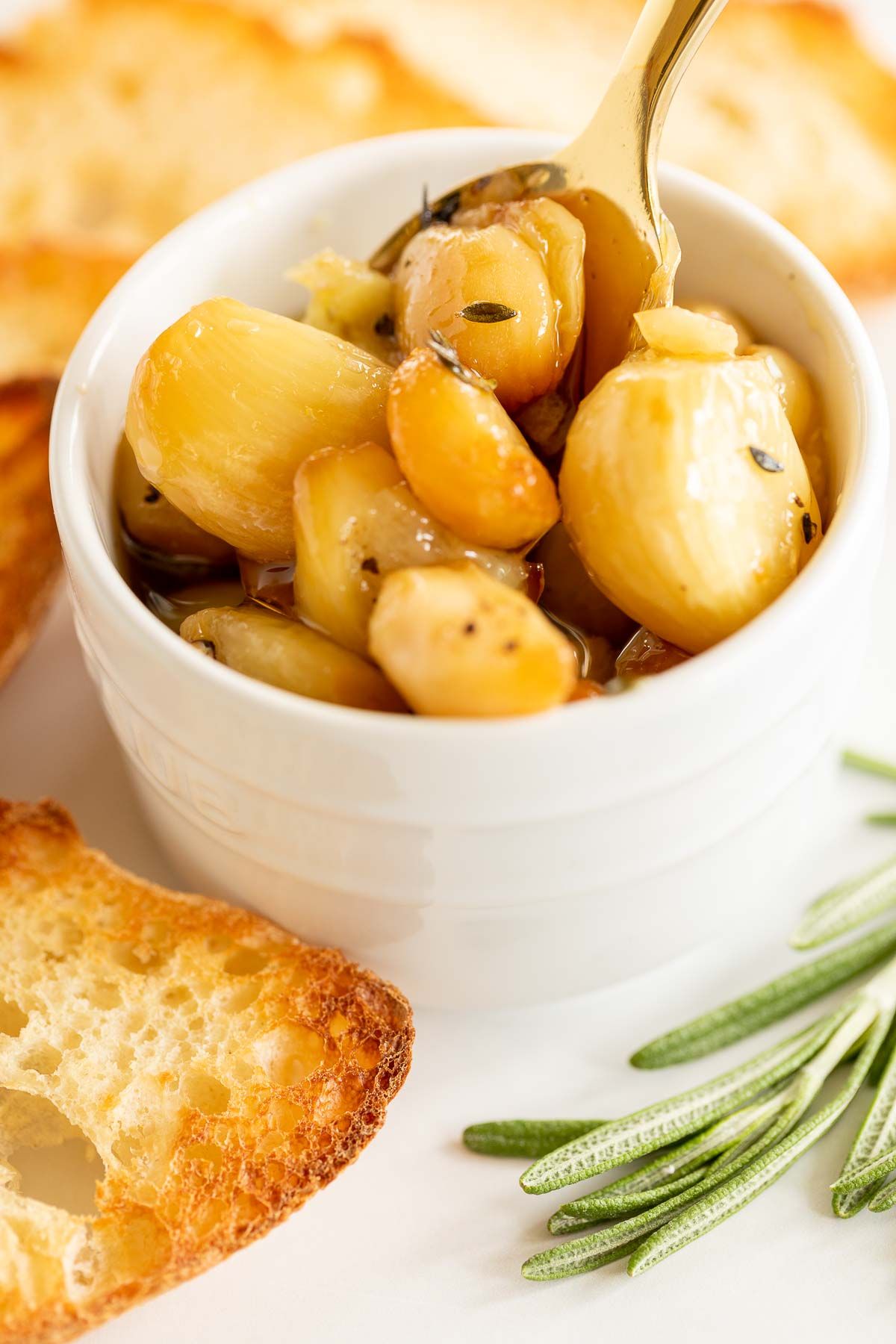 A white bowl full of garlic confit, surrounded by crostini and fresh rosemary. 