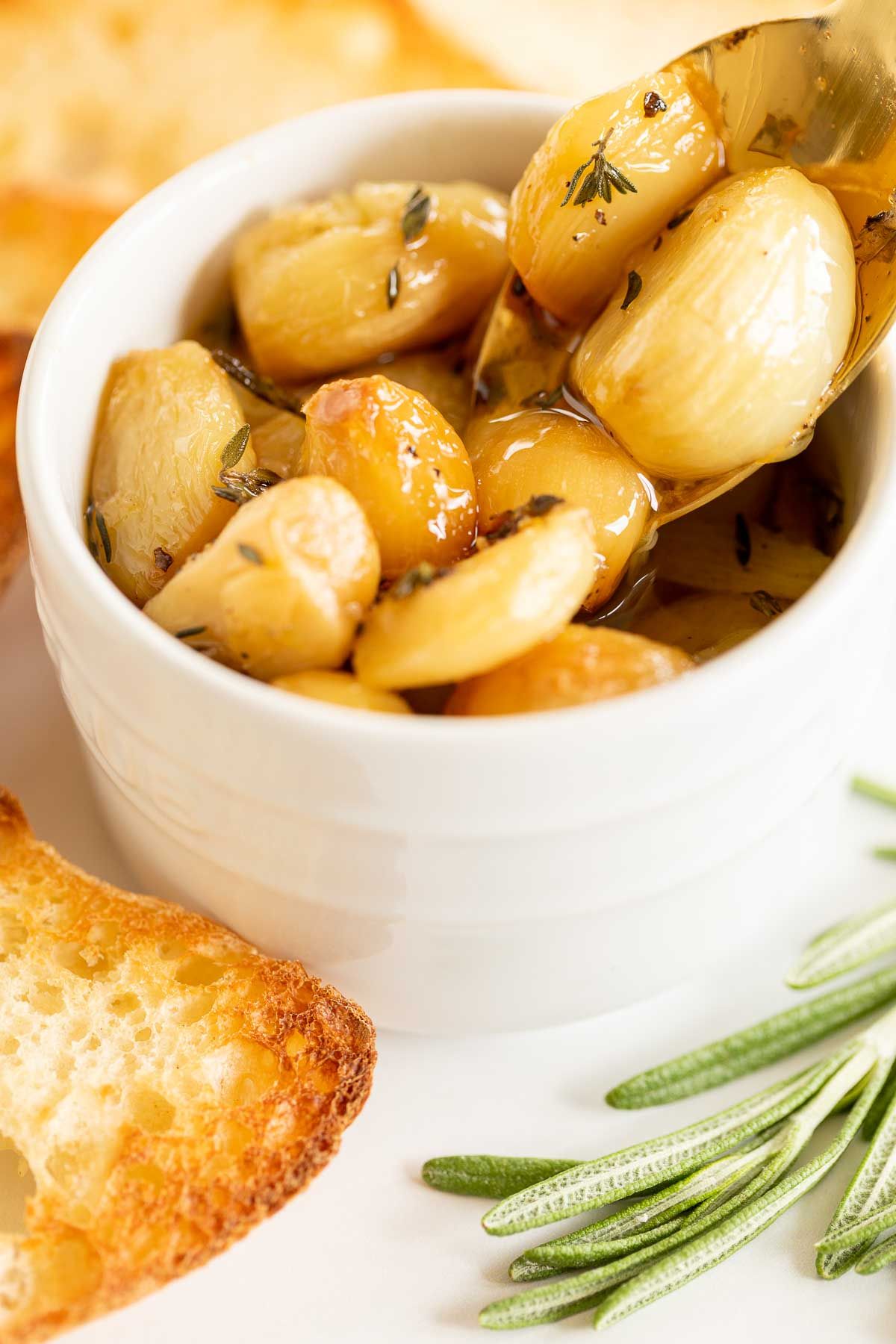 A white bowl full of garlic confit, surrounded by crostini and fresh rosemary.