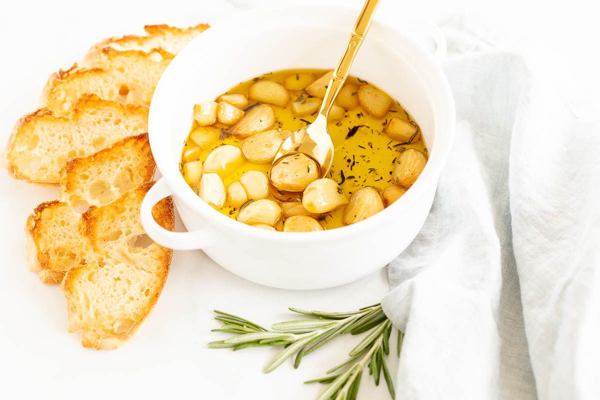 A white bowl full of garlic confit, surrounded by crostini and fresh rosemary.