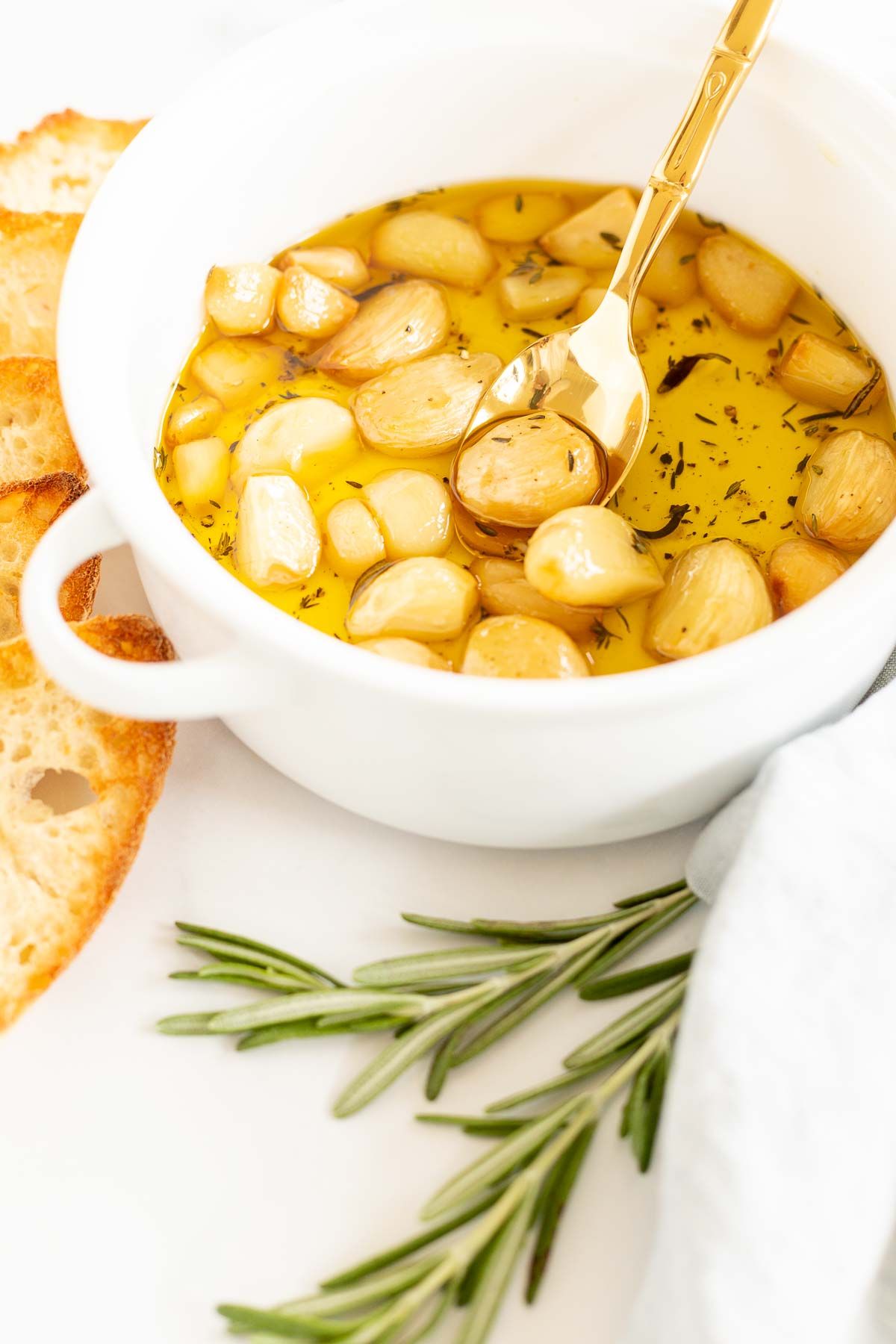 A white bowl full of garlic confit, surrounded by crostini and fresh rosemary.