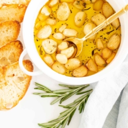 A white bowl full of garlic confit, surrounded by crostini and fresh rosemary.