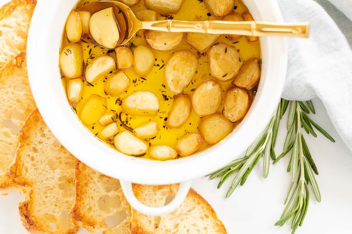 A white bowl full of garlic confit, surrounded by crostini and fresh rosemary. 