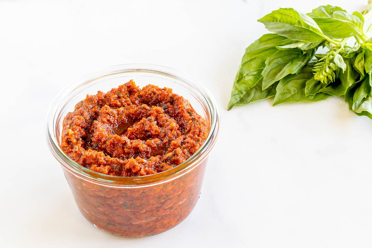 A small white bowl of pesto rosso, placed next to basil leaves, placed on a marble countertop.