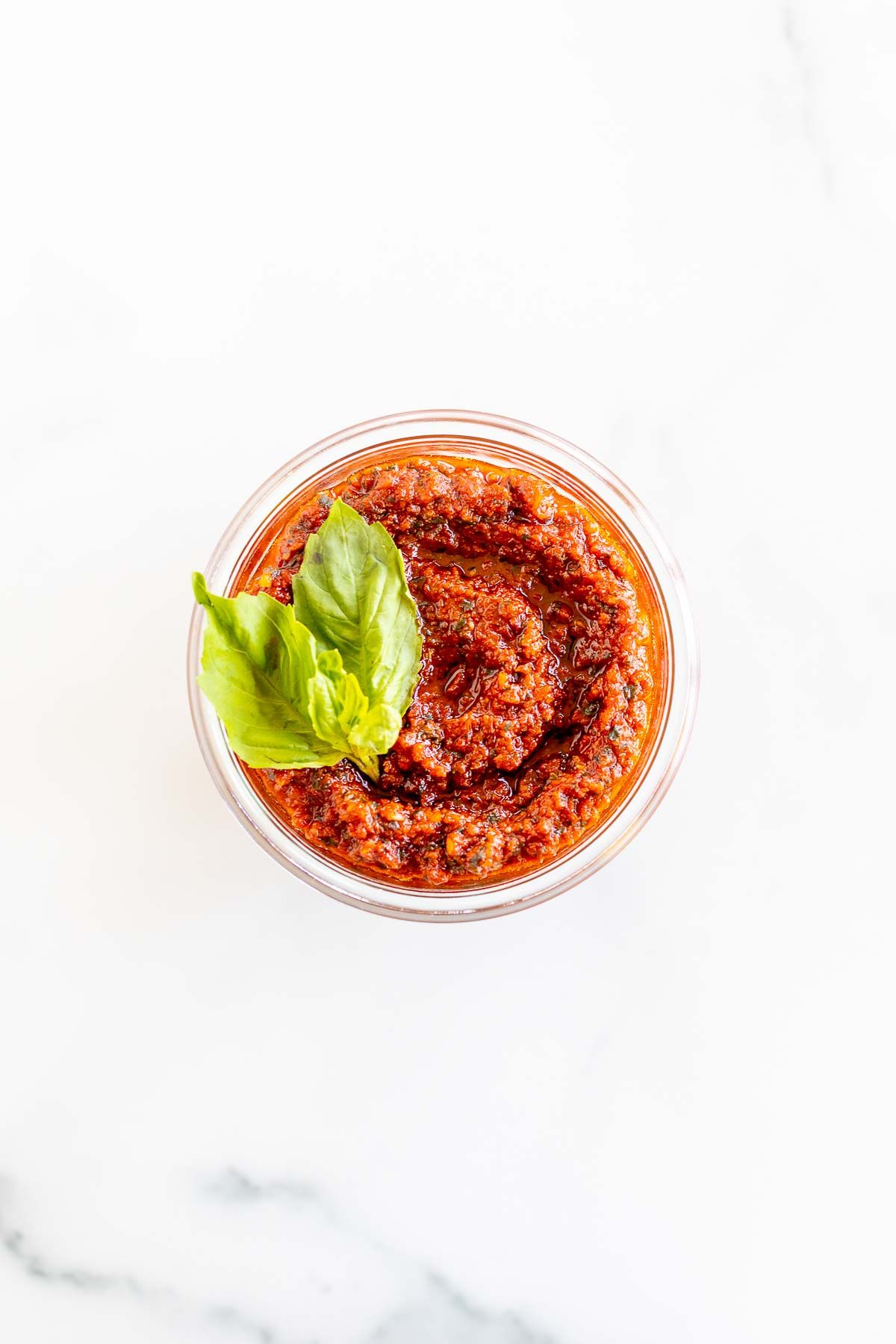 A small white bowl of pesto rosso, garnished with fresh basil leaves, placed on a marble countertop.