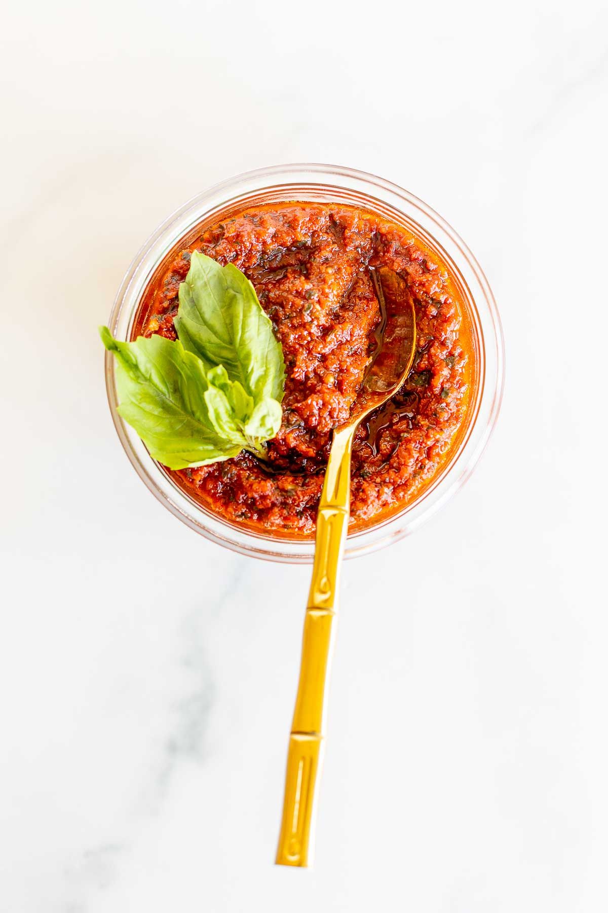 A small white bowl of pesto rosso, garnished with fresh basil leaves, placed on a marble countertop. Gold spoon tops the bowl.