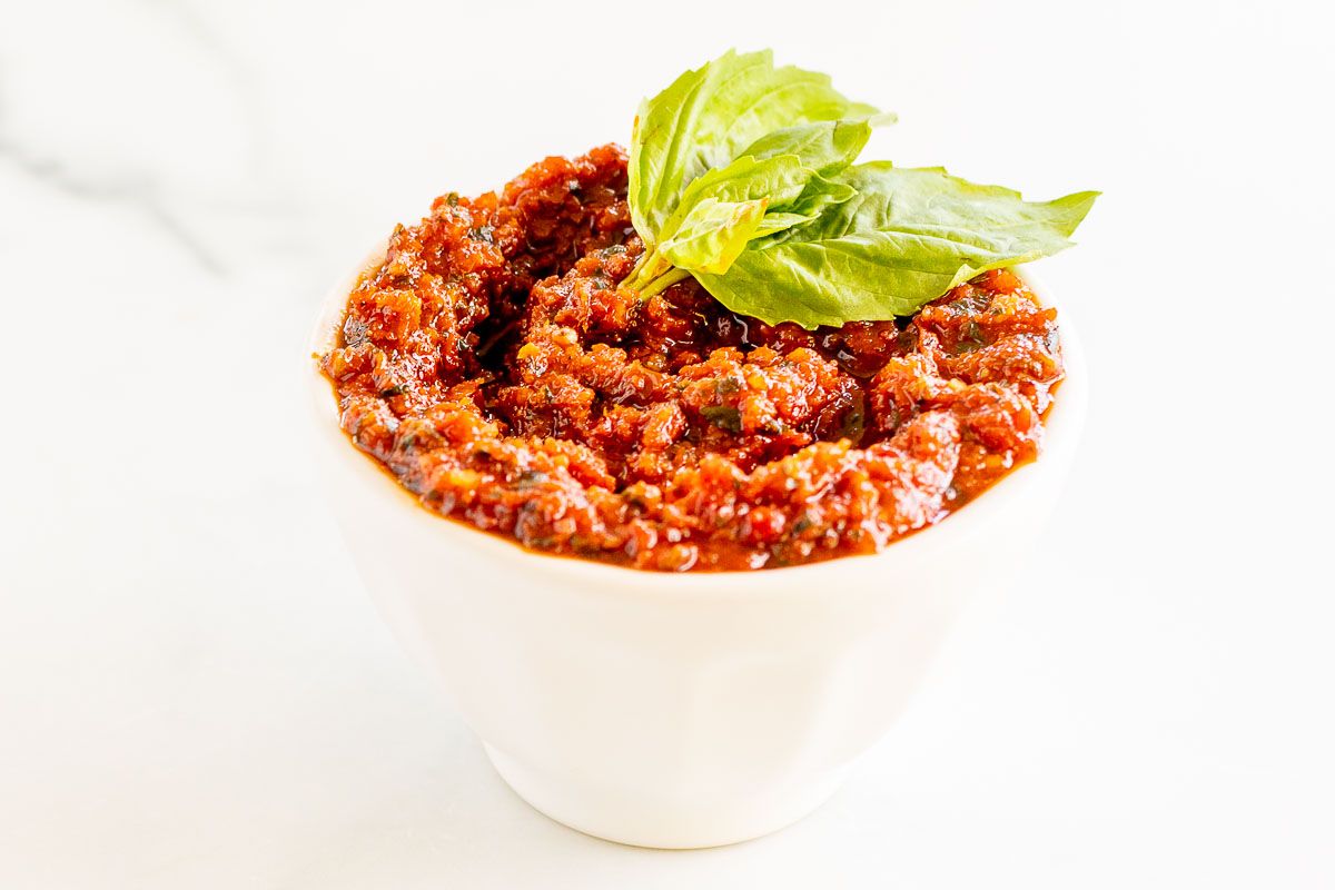A small white bowl of pesto rosso, garnished with fresh basil leaves, placed on a marble countertop.
