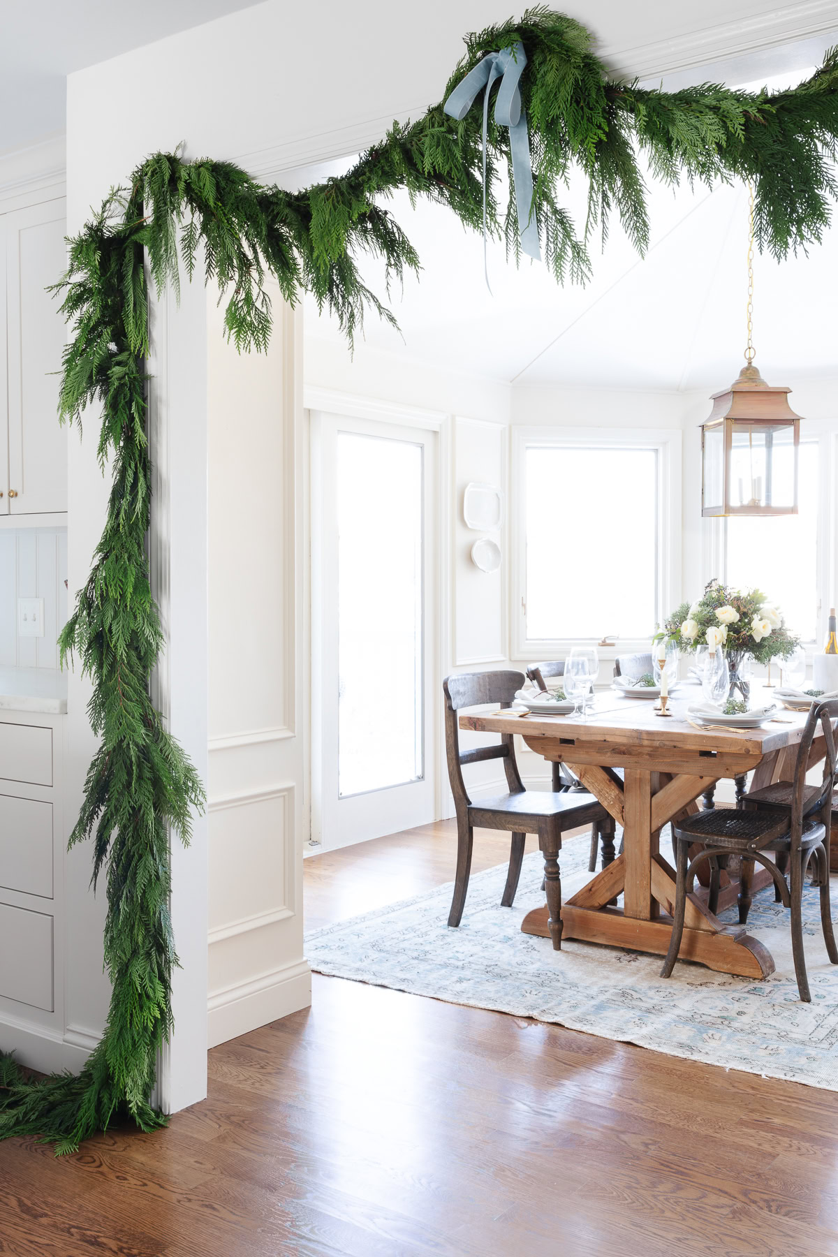 A dining room with a wooden table set for a meal, featuring a large cedar garland draped over a doorway and natural light streaming through the windows.