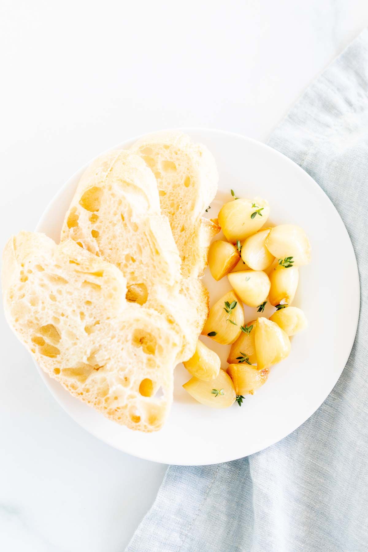 A white plate with sliced French bread and cloves of roasted garlic.