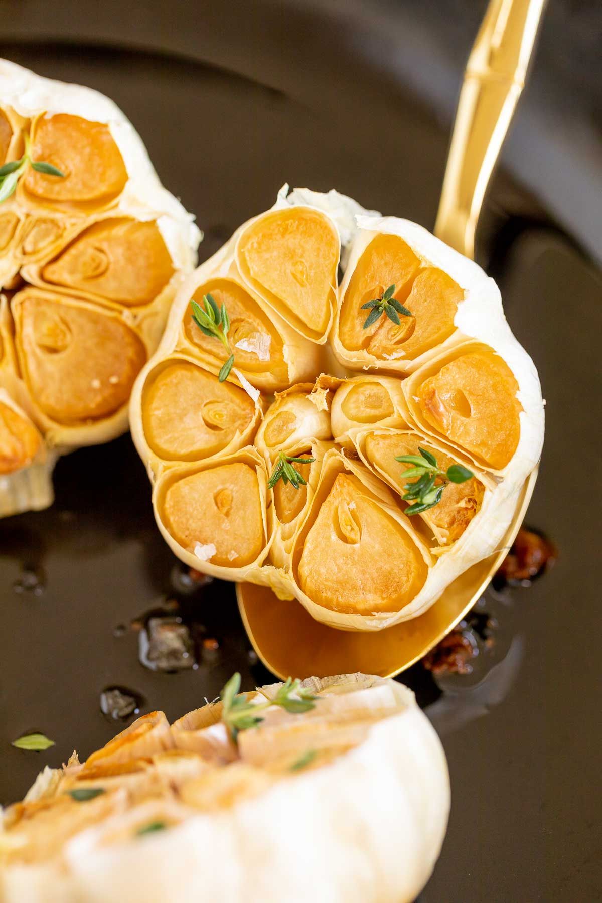 A gold spoon lifting up a head of roasted garlic from a cast iron pan