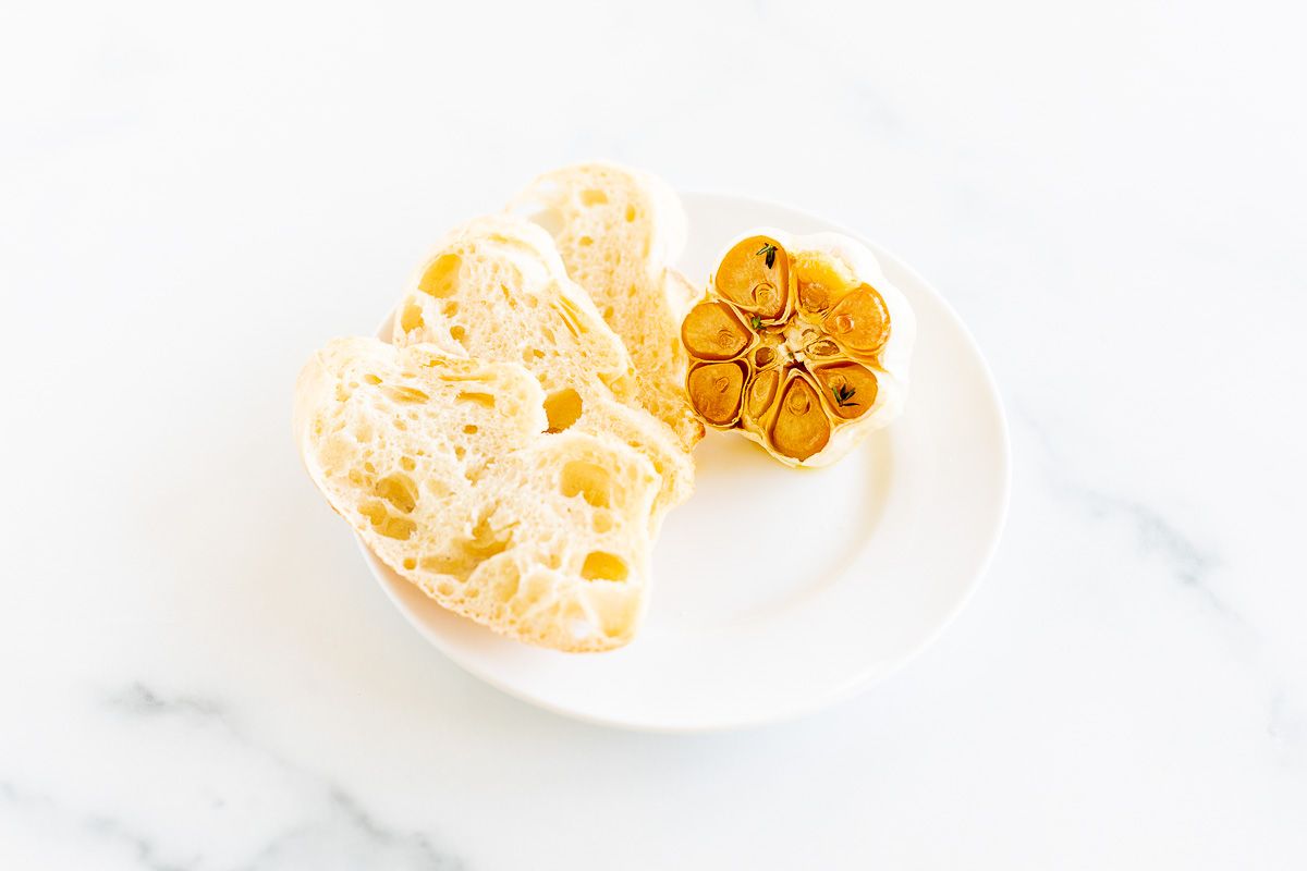 A white plate holes sliced French bread and a head of roasted garlic.