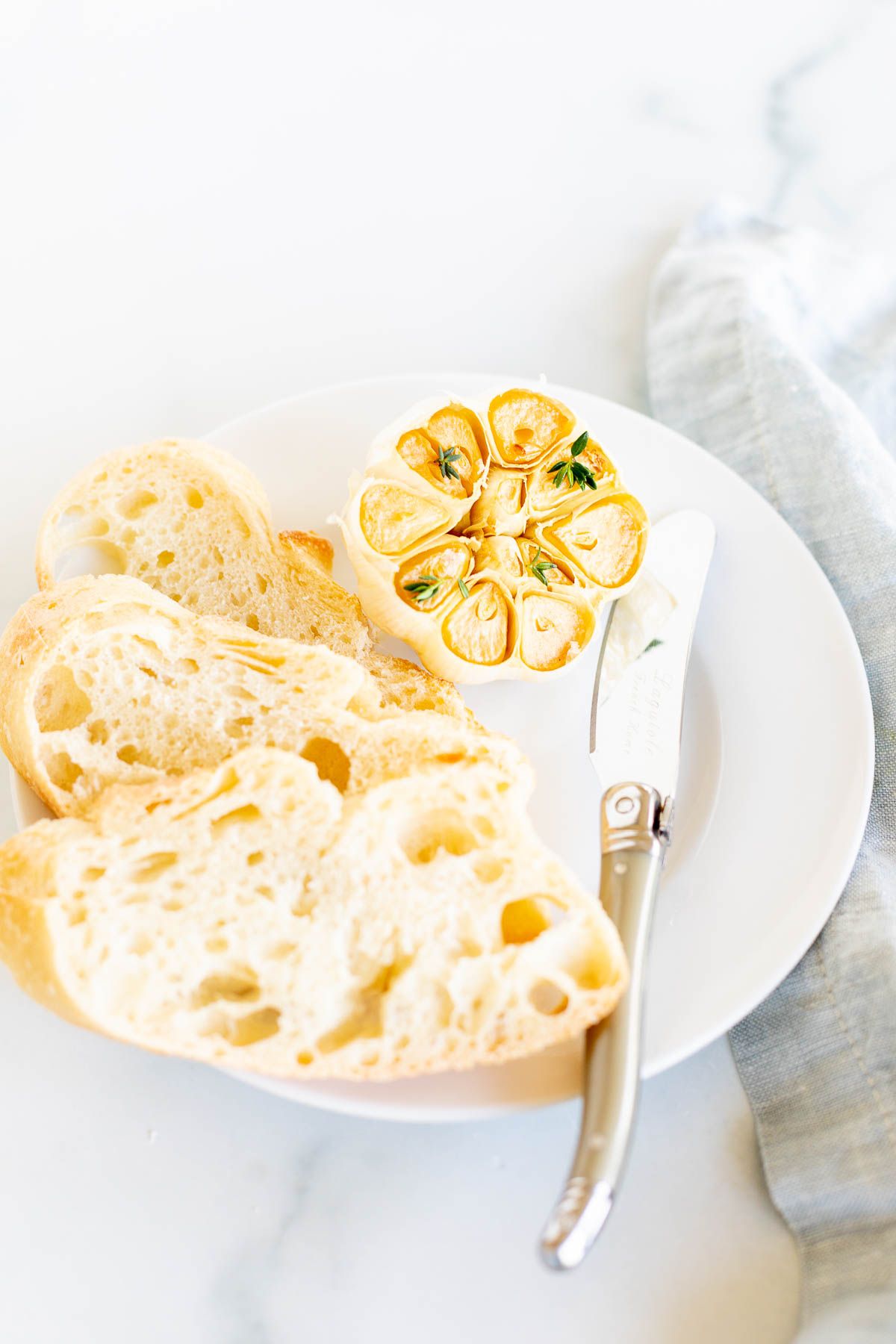 A white plate holes sliced French bread and a head of roasted garlic.