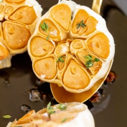 A gold spoon lifting up a head of roasted garlic from a cast iron pan