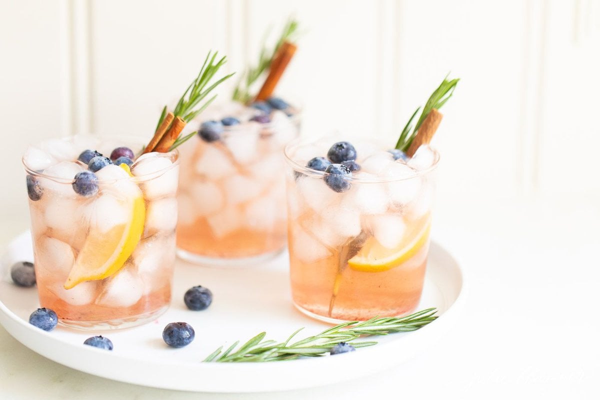 Three Smash cocktails garnished with rosemary, blueberries, oranges, and cinnamon sticks all on a white plate. 