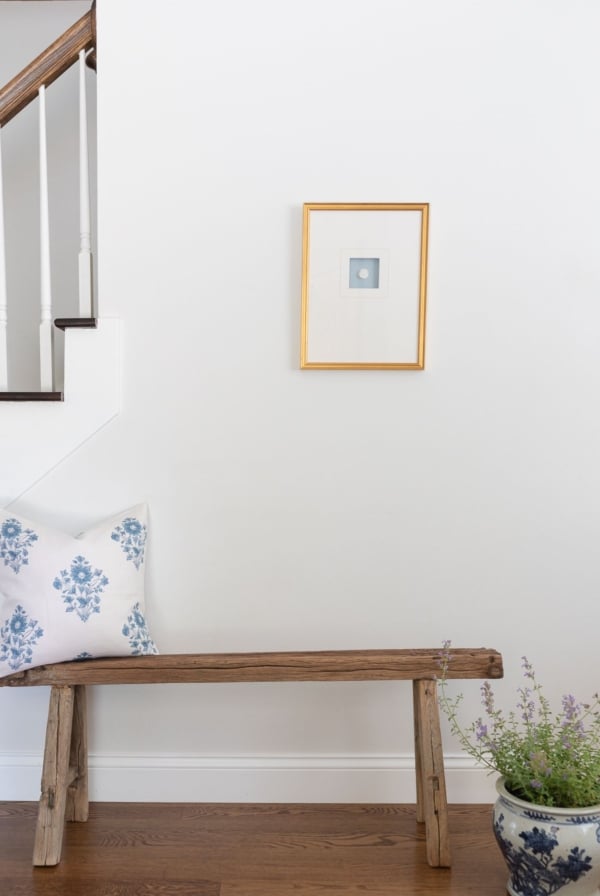 A gold framed intaglio in a white entryway of a home, wooden bench below.