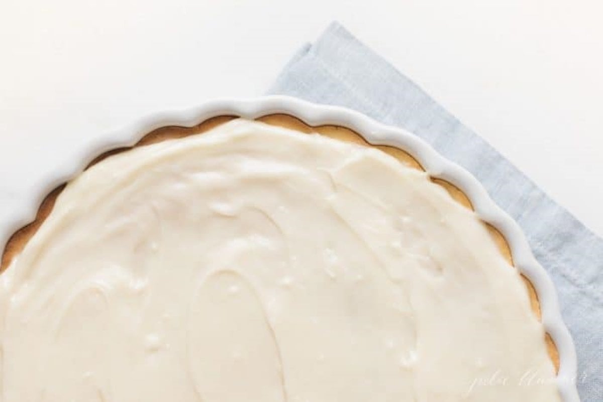 A fruit pizza with fruit pizzing icing in a white dish, blue towel in background.