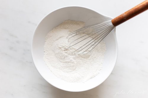 A white bowl containing flour and peanut butter with a metal whisk partially immersed, set on a white marble surface.