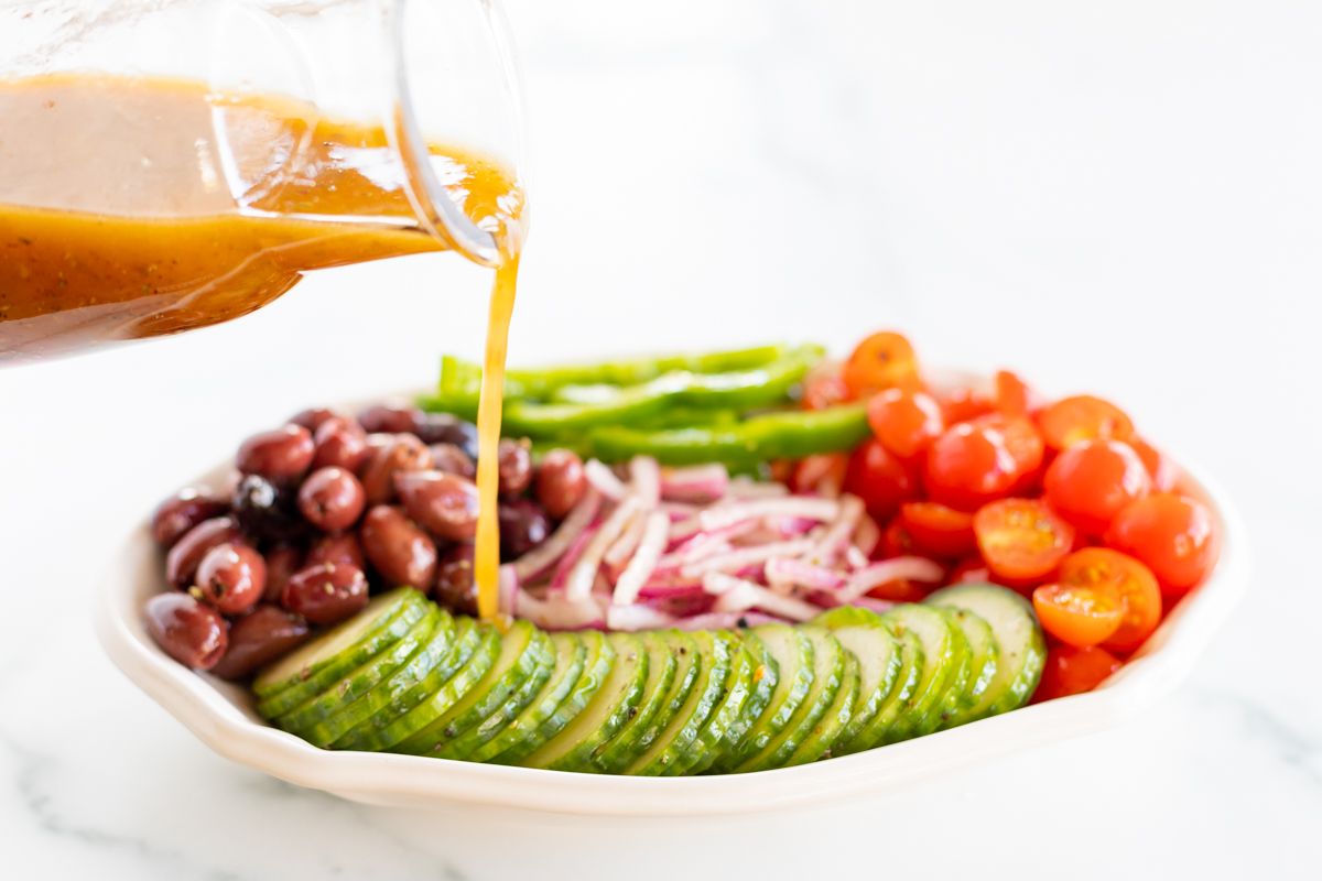 A small glass jar of Greek salad dressing pouring over a colorful Greek Salad on a white platter.