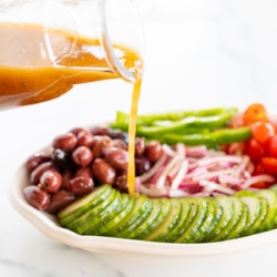 A small glass jar of Greek salad dressing pouring over a colorful Greek Salad on a white platter.