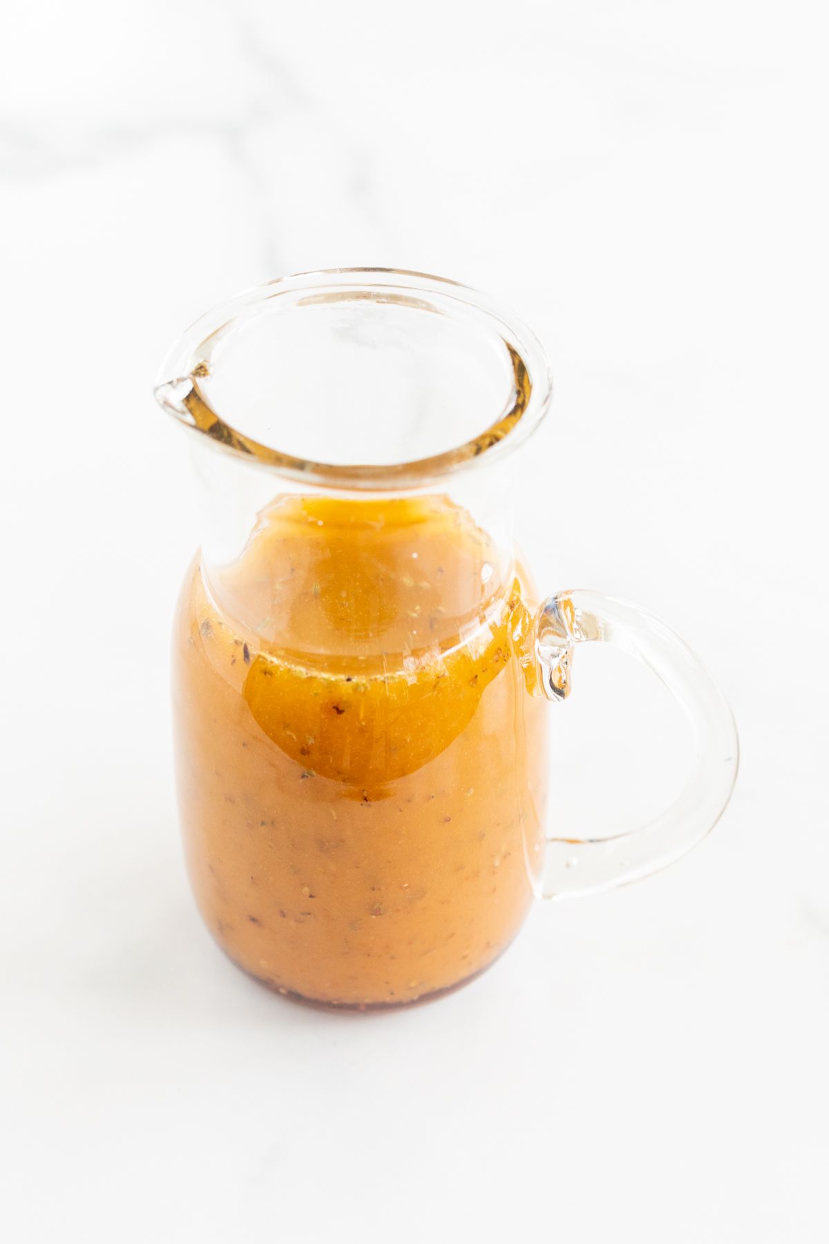 A small glass pitcher full of homemade Greek Salad dressing, on a white countertop.